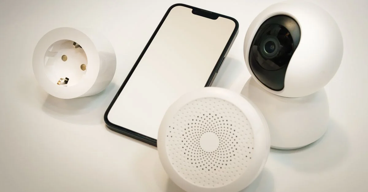 Gadgets de maison intelligente incluant une caméra de sécurité, un haut-parleur et un smartphone affichés sur une surface blanche.